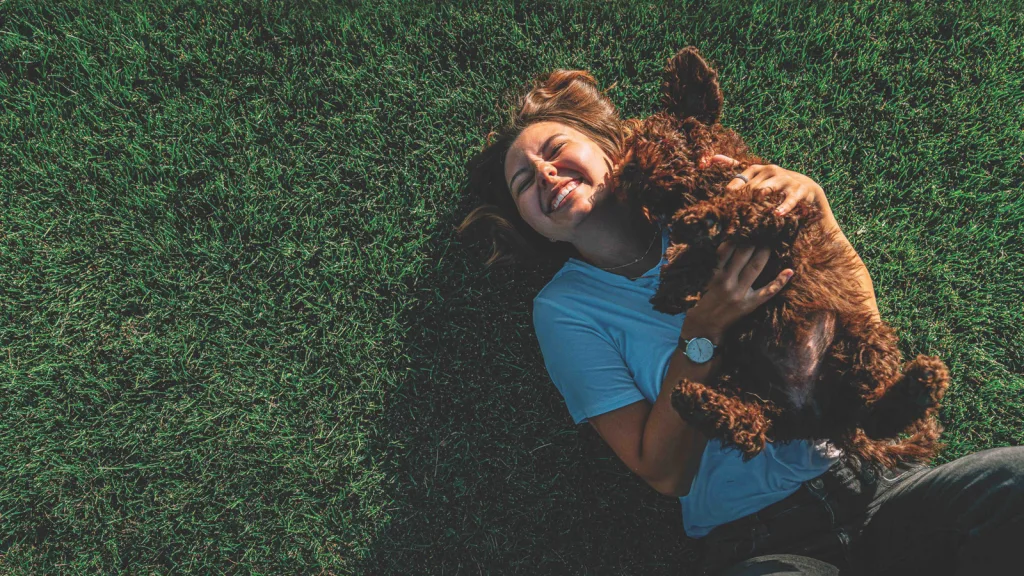 Woman lying in grass with her chestnut-colored poodle on her chest licking her face. The woman is smiling and holding her dog. She is wearing a watch on her right arm and wearing a blue t-shirt and black denim jeans. New pet-friendly apartment homes for rent with private fenced backyards in Arizona, Texas, Florida, and North Carolina with pool, pickleball, dog park, gym, clubhouse and gated community. *No breed or weight restrictions. Yardly